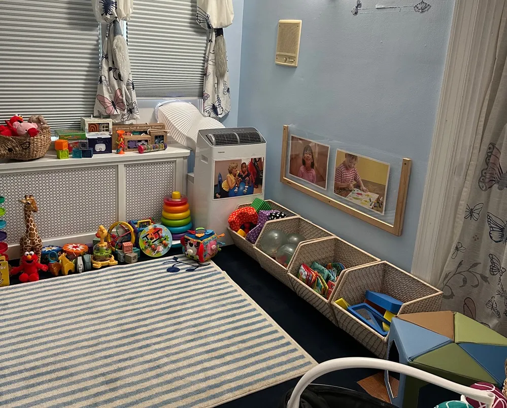 A child care playroom is filled with toys, baskets, blocks and a striped rug, with children's photos displayed along a light blue wall.