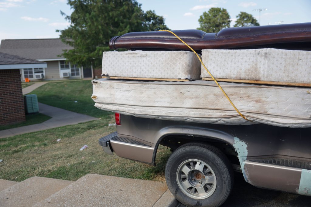 Several mattresses are stacked in a truck with an apartment complex in the background.