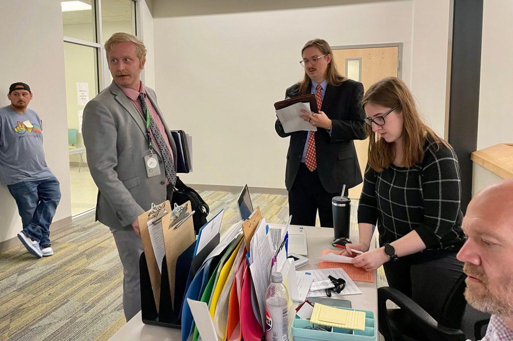 A group of people sit and stand around a table with paperwork piled atop it.
