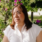 Cynthia Villarreal, wearing a white blouse, stands outdoors among flowering bushes and trees, looking off to the side in the sunlight.