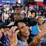 Keisha Lance Bottoms smiles while taking a selfie with a supporter at a crowded campaign rally, with cameras and attendees gathered behind them.