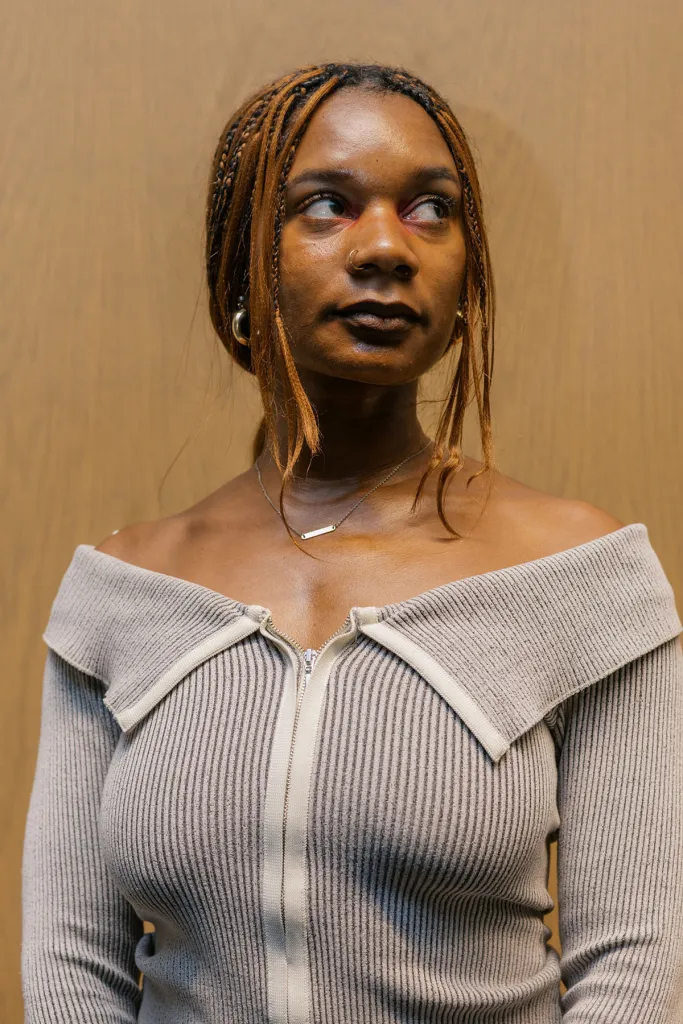A young woman with blonde-highlighted braids, a nose ring and an off-the-shoulder beige sweater looks to her right while posing against a tan wall.