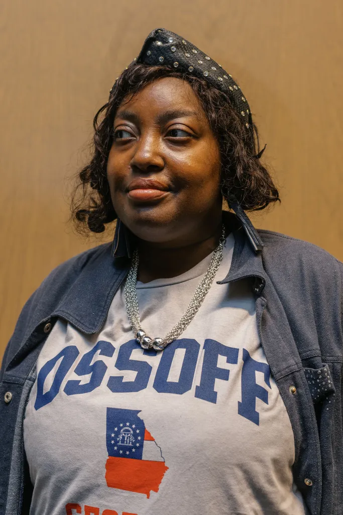 A woman wearing a studded black hat, layered silver necklaces and an Ossoff T-shirt looks to her right against a tan wall.