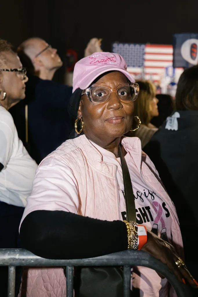 A woman in a pink "Fight" baseball cap, clear-framed glasses and a pink quilted vest stands behind a metal barricade at a crowded campaign rally.