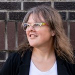 A person with shoulder-length light brown hair and glasses smiles while looking to the left of the frame. They wear a black blazer over a white shirt and stand in front of a dark red and black brick wall.