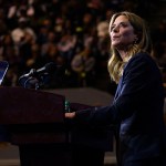 Amy Acton stands at a podium with two microphones, looking out toward the audience at an indoor campaign event. She wears a dark blazer, with a crowd of supporters visible in the dimly lit background.