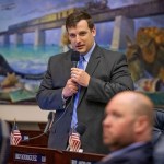 Mike Beltran, wearing a gray suit and blue striped tie, speaks into a microphone in a legislative chamber.