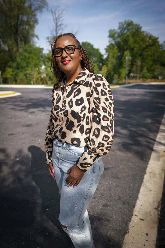 Darnese Foster wearing black-framed glasses, a leopard print blouse and jeans stands outdoors on a sunny residential street.