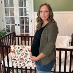 A pregnant woman stands beside a wooden crib in a nursery, resting her hands on her belly and looking toward the camera.