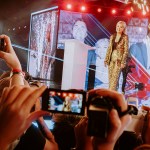 Erika Kirk in a gold sequined suit stands onstage surrounded by pyrotechnics and large video screens as audience members hold up their phones to photograph her.