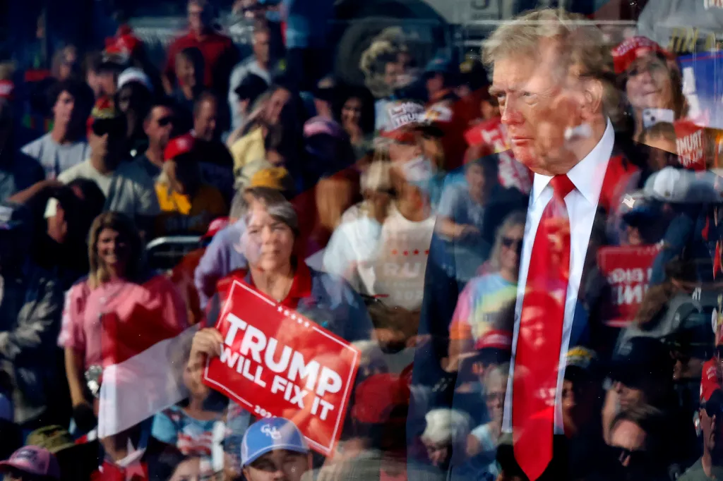 Donald Trump is seen through a reflective surface, with a crowd of supporters and a red "Trump Will Fix It" sign visible around his image.