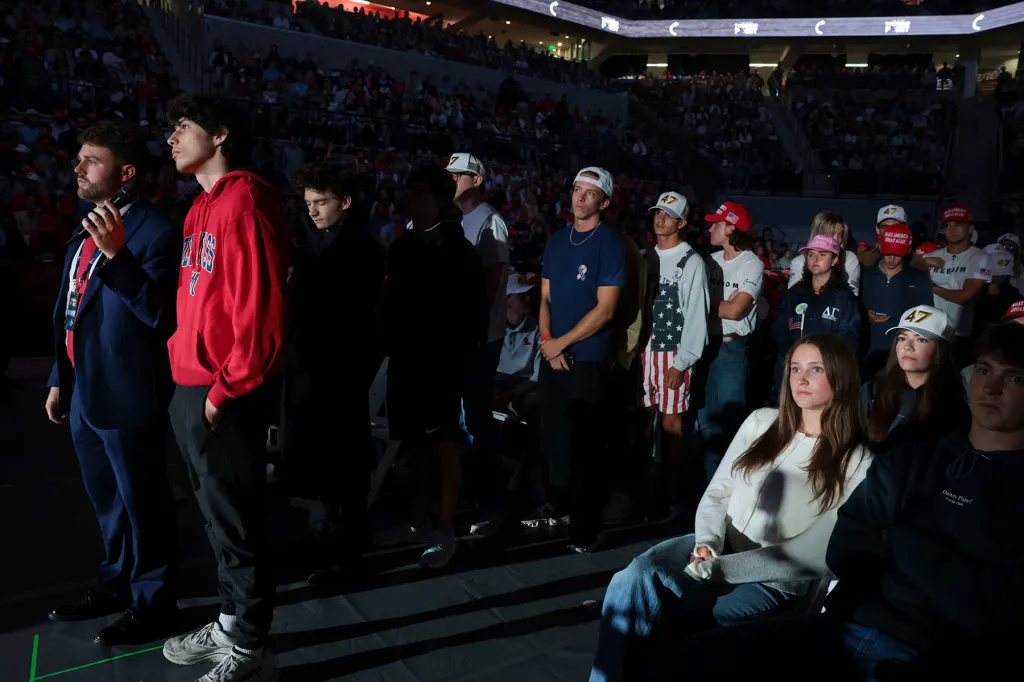 Young attendees wearing Turning Point USA merchandise stand and sit in a dimly lit arena, illuminated by stage lighting.