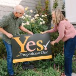 Steve and Lisa smile at each other as they place a “Yes on Proposition One” sign in their front garden.