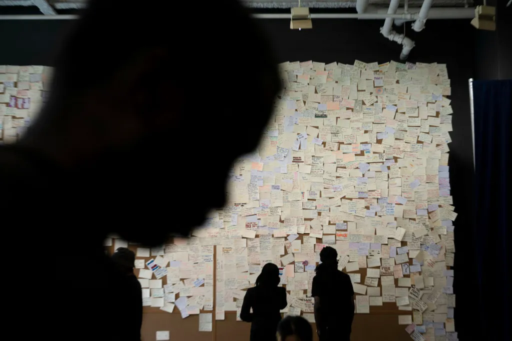 Visitors stand before a large wall covered in hundreds of handwritten notes at an exhibition about Donald Trump and Jeffrey Epstein. A person's dark silhouette fills much of the foreground.
