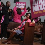 Angie Nixon laughs while seated in a church pew at a campaign event, wearing a bright pink blazer. Supporters around her hold pink Angie Nixon for U.S. Senate signs.