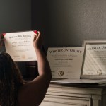 A Black woman with her back turned to the camera places her college degrees atop a dresser