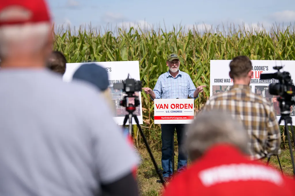 Rep. Derrick Van Orden speaks at a press conference in front of a cornfield as journalists with cameras look on.