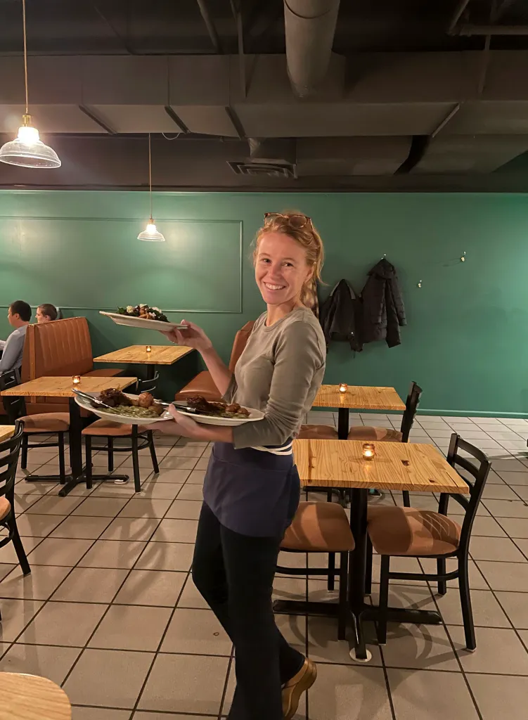 Rebecca Cooke smiles while carrying plates of food inside a restaurant.