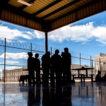 Silhouettes of people gather around picnic tables beneath a covered outdoor area at a detention facility. Chain-link fences topped with razor wire and large buildings surround the space.