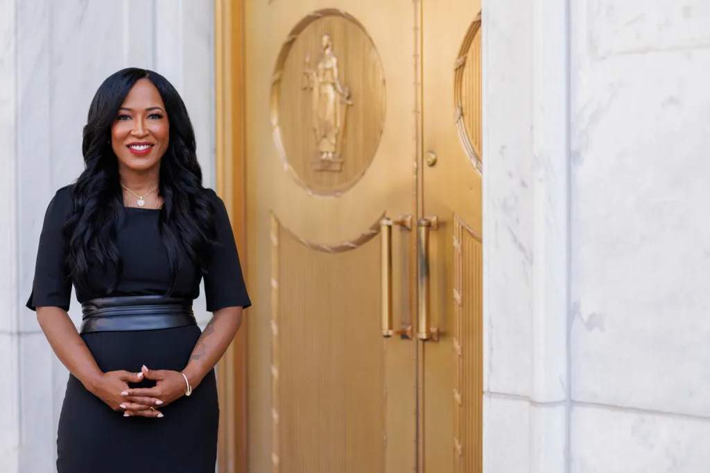 Miller stands alongside a white building and gold door, wearing a black dress and smiling at the camera