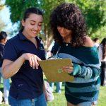 A person helps another person fill out a form outside in a grassy area surrounded by other people.