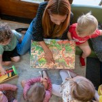 Overhead view of a woman reading a colorful dinosaur book to several young children gathered around her in a childcare setting.