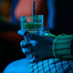 Close-up of a person wearing rings holding a clear drink with a lime wedge and straw in a dimly lit bar, illuminated by blue and green light.
