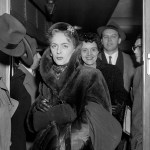 Christine Jorgensen stands in a doorway wearing a dark fur coat, scarf and gloves, holding a small handbag. Several people stand behind her, including a smiling woman and a man in a hat.