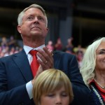 Sen. Roger Marshall in a navy suit and red tie applauds beside a smiling woman with long blonde hair at an indoor event. Two children are visible in the foreground.