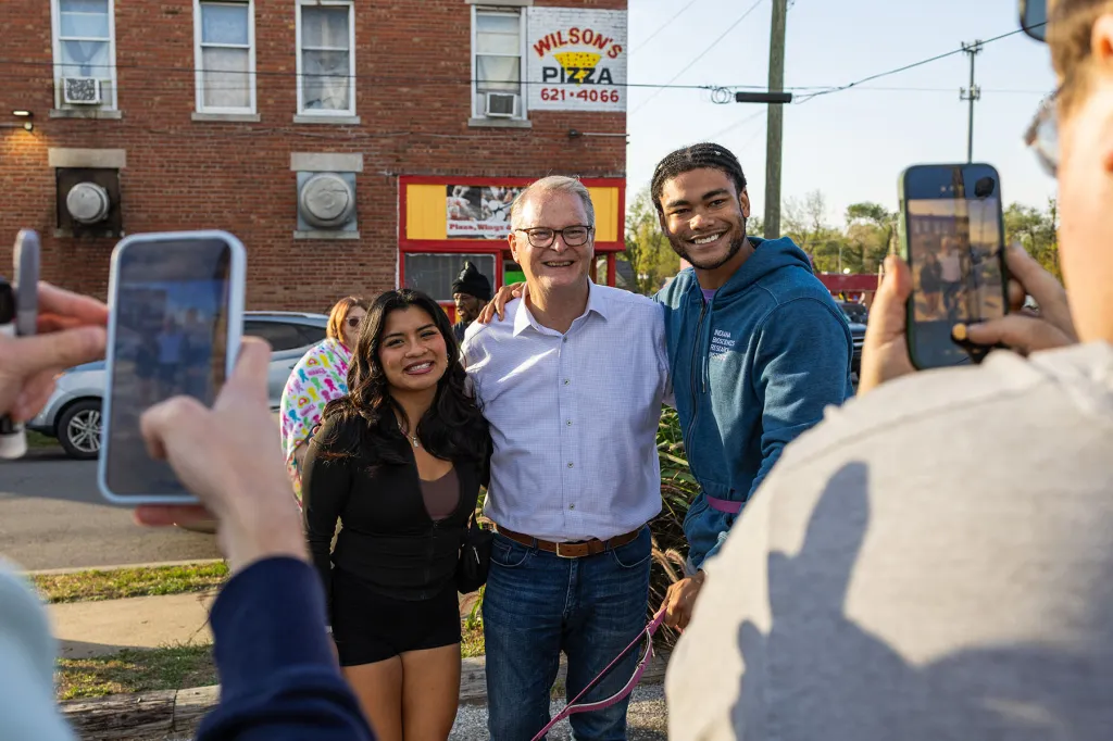 Adam Hamilton poses with supporters outside a brick building as people in the foreground photograph them with smartphones.