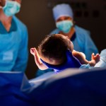 A newborn is held up by a gloved medical worker in an operating room as masked staff look on in the background.