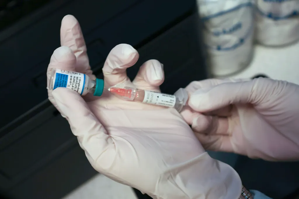 Gloved hands draw a measles vaccine dose into a syringe from a vial.