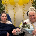Melissa Napolitan (left) and her mother, Cindy, sit side by side holding hands. Cindy holds a bouquet of flowers, and yellow balloons and hanging greenery decorate the wall behind them.