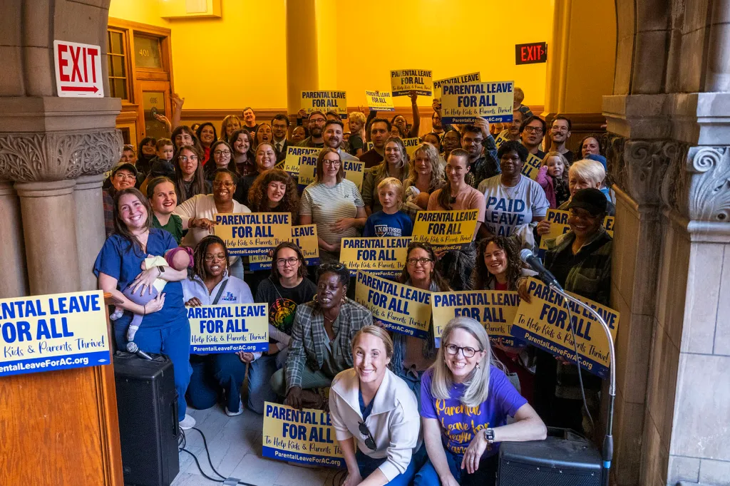 A large group of people, including nurses in scrubs and parents holding young children, pose on a staircase inside a government building. Many hold yellow and blue signs reading "Parental leave for all."