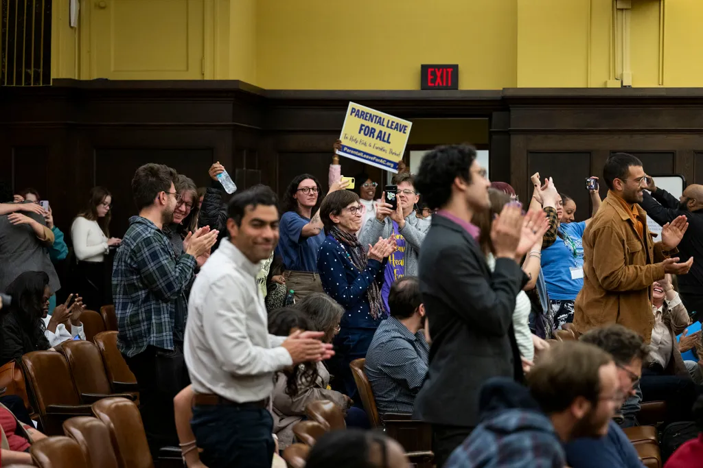 People stand and applaud inside a crowded council chamber. Several raise their hands, hold up phones or display yellow and blue "Parental leave for all" signs.