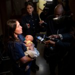 A woman in navy blue scrubs holds a baby in a yellow outfit as reporters surround her with microphones, cameras and recording devices in a dimly lit hallway.