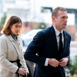 Max Miller, wearing a navy suit and tie, walks alongside Emily Moreno, dressed in a cream-colored coat, outside a building in Washington.