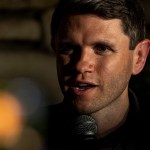 James Talarico speaks into a microphone, his face partially illuminated by warm light against a dark, blurred background.